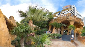 A hotel entrance with a natural rock facade, palm trees, and a sign indicating 'AMIGOS'. - Magic Aqua Rock Gardens (Benidorm)