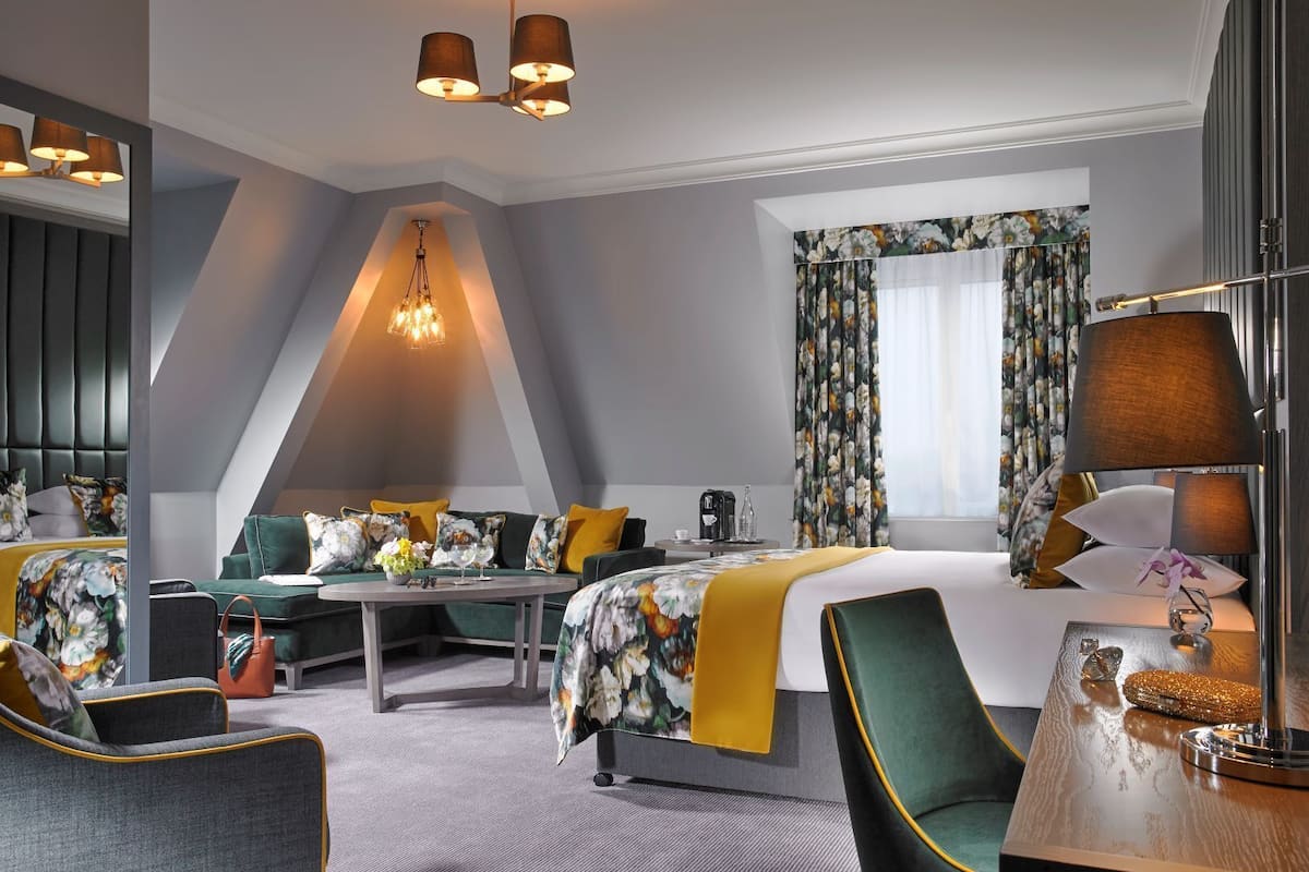 Ein modernes Hotelzimmer mit Bett, Sofa, Schreibtisch und Stuhl. Ein Fenster mit Vorhängen, eine Kandelaber und eine Lampe sind ebenfalls vorhanden.