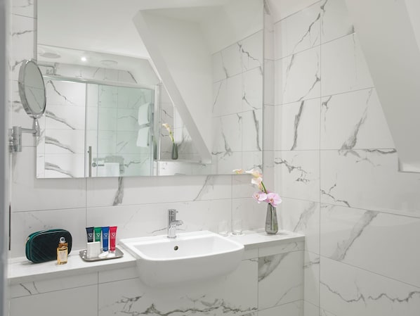 Baño con lavamanos blanco, espejo y azulejos de mármol. Hay artículos de aseo en la encimera y un florero con flores.