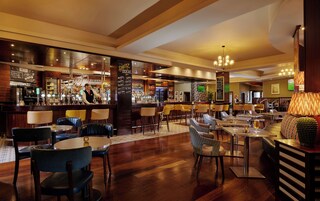 Zona de bar con un barman, diversas opciones de asientos que incluyen taburetes y mesas, y una variedad de equipo de bar.