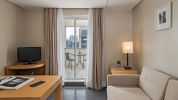 Ett hotellrum med en platt-TV, ett skrivbord i trä, en soffa, ett litet bord med en lampa och en balkong med utsikt över byggnaderna.