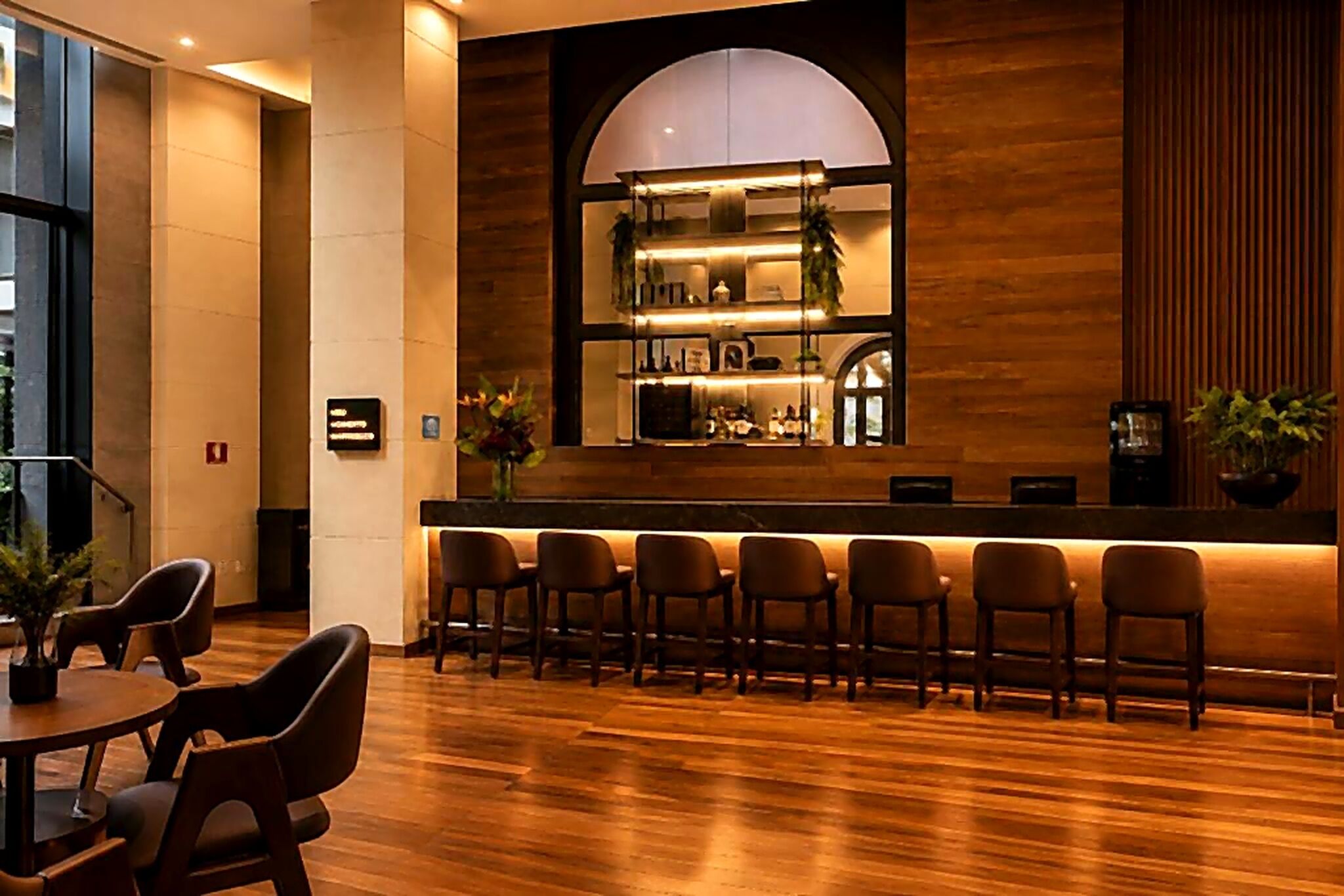 Een bar met een lange toonbank, meerdere stoelen en een houten wand met een groot rondboogvenster.