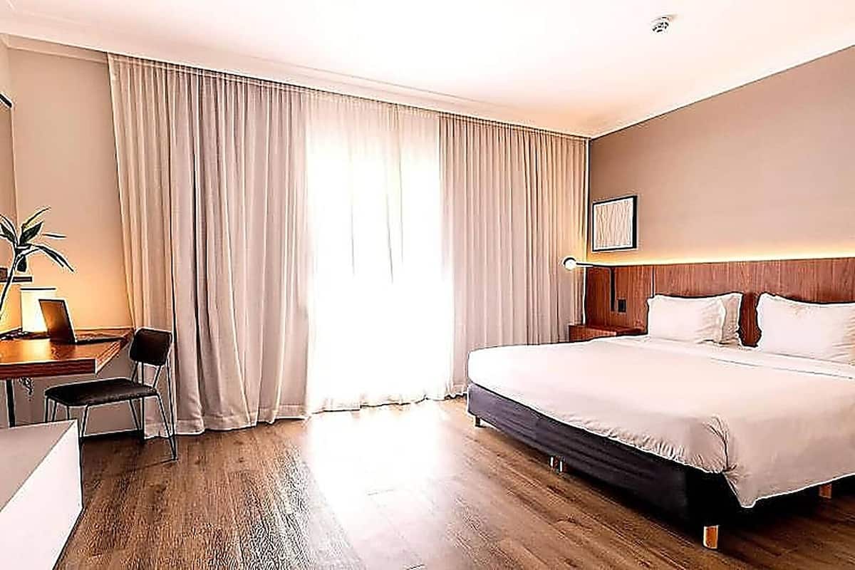 Een hotelkamer met een bed, een bureau met stoel, een lamp en een groot raam met gordijnen.