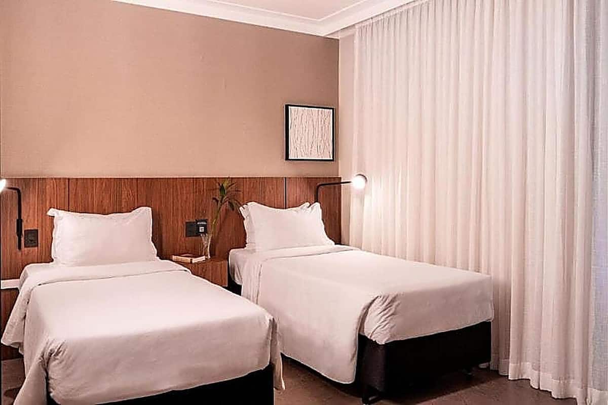 Een hotelkamer met twee bedden, een nachtkastje met een lamp, een ingelijst schilderij aan de muur en een groot raam met gordijnen.