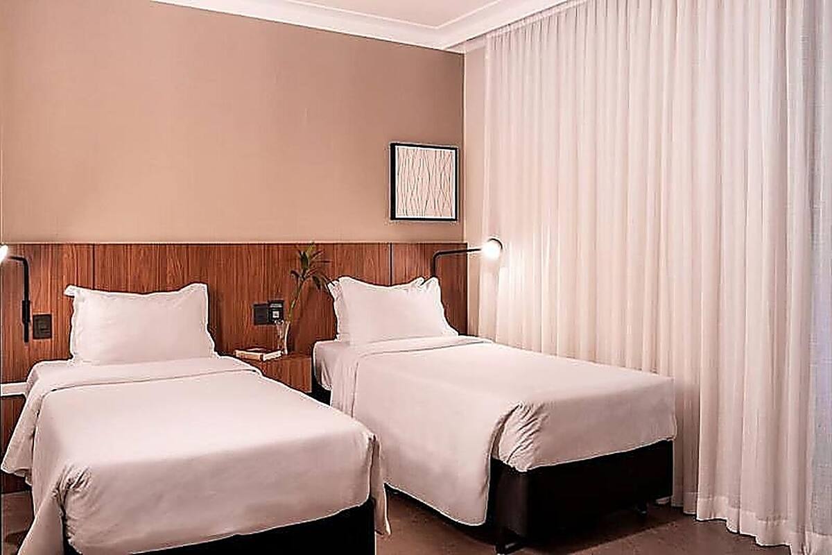 Een hotelkamer met twee bedden, een houten hoofdbord, een nachtkastje met een lamp en een groot raam met gordijnen.