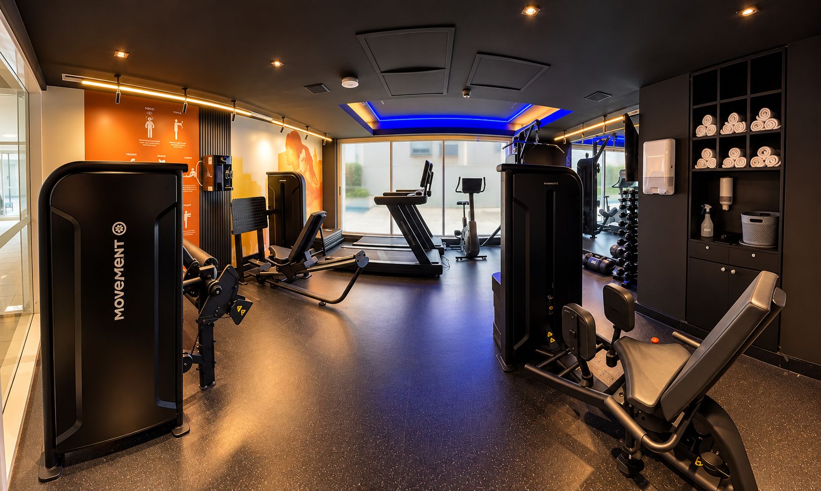 Ett gym med olika typer av träningsutrustning, inklusive löpband och styrketräningsmaskiner, samt en vägg med en handduksställning.