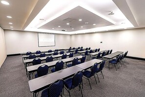 Een congreszaal met rijen tafels en stoelen, een scherm voor een projector en een plafond met inbouwspots.