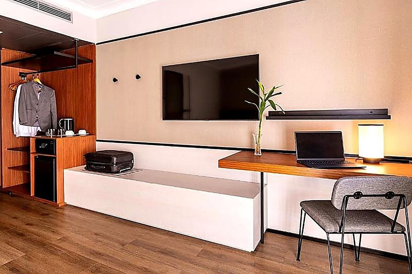 Quarto de hotel com guarda-roupa de madeira, secretária com um laptop, uma cadeira, televisão e uma pequena planta.