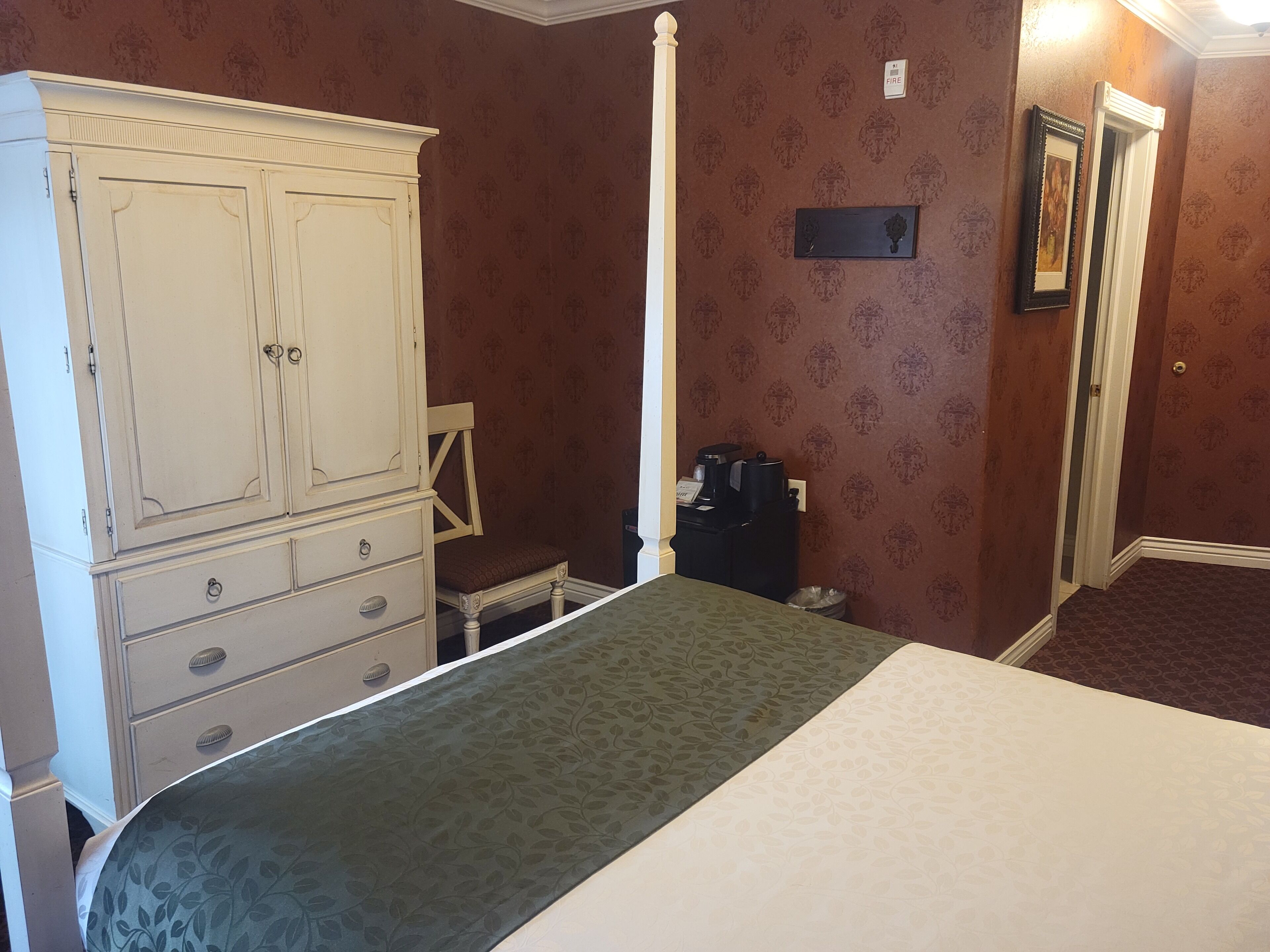 Quarto com um guarda-roupa grande e branco, uma cama com colcha verde, uma cadeira e uma mesa pequena com cafeteira.