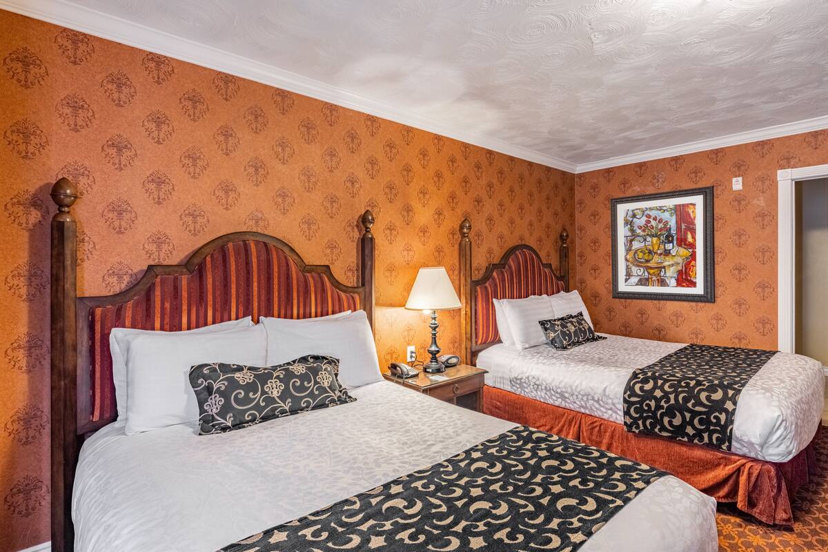 Habitación de hotel con dos camas, una mesita de noche con lámpara, un cuadro enmarcado en la pared y papel tapiz estampado.