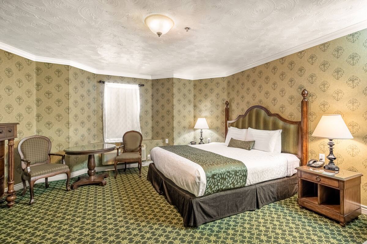 Una habitación de hotel con una cama grande, dos sillas, una mesita y una mesita de noche con lámpara.