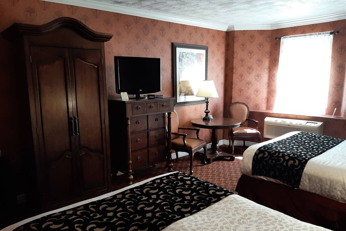 Habitación de hotel con dos camas, un aparador, televisor, silla, mesa y ventana con cortinas.