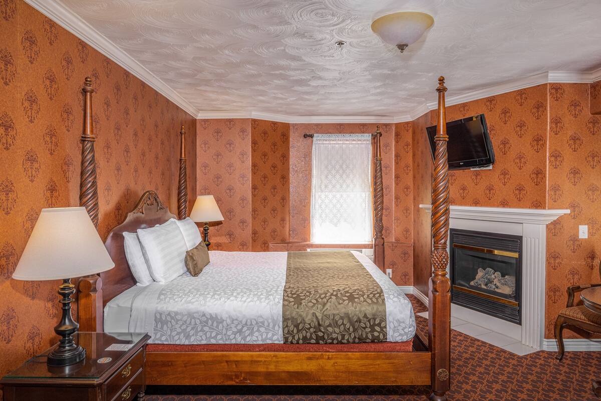 Una cama con dosel y tablero de madera, una mesita de noche con lámpara, chimenea y televisión fijada en la pared.