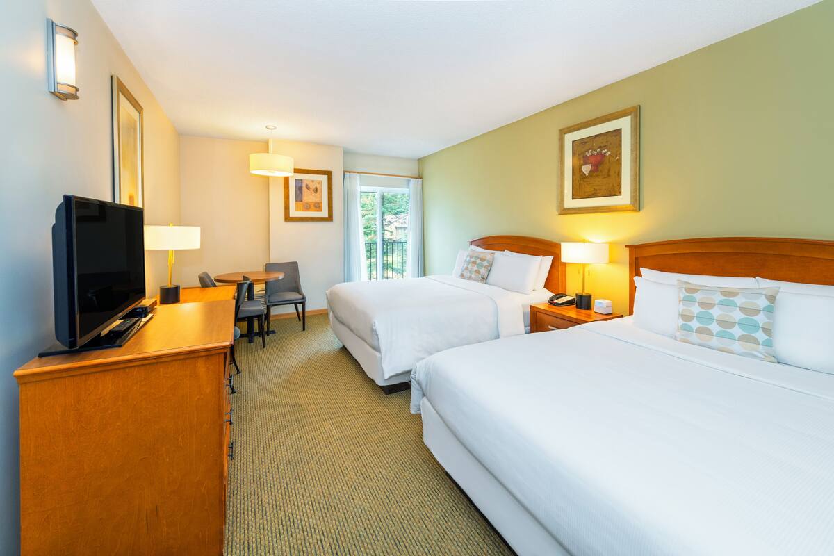 A hotel room with two beds, a desk with a TV, a chair, and a window with a view of greenery.