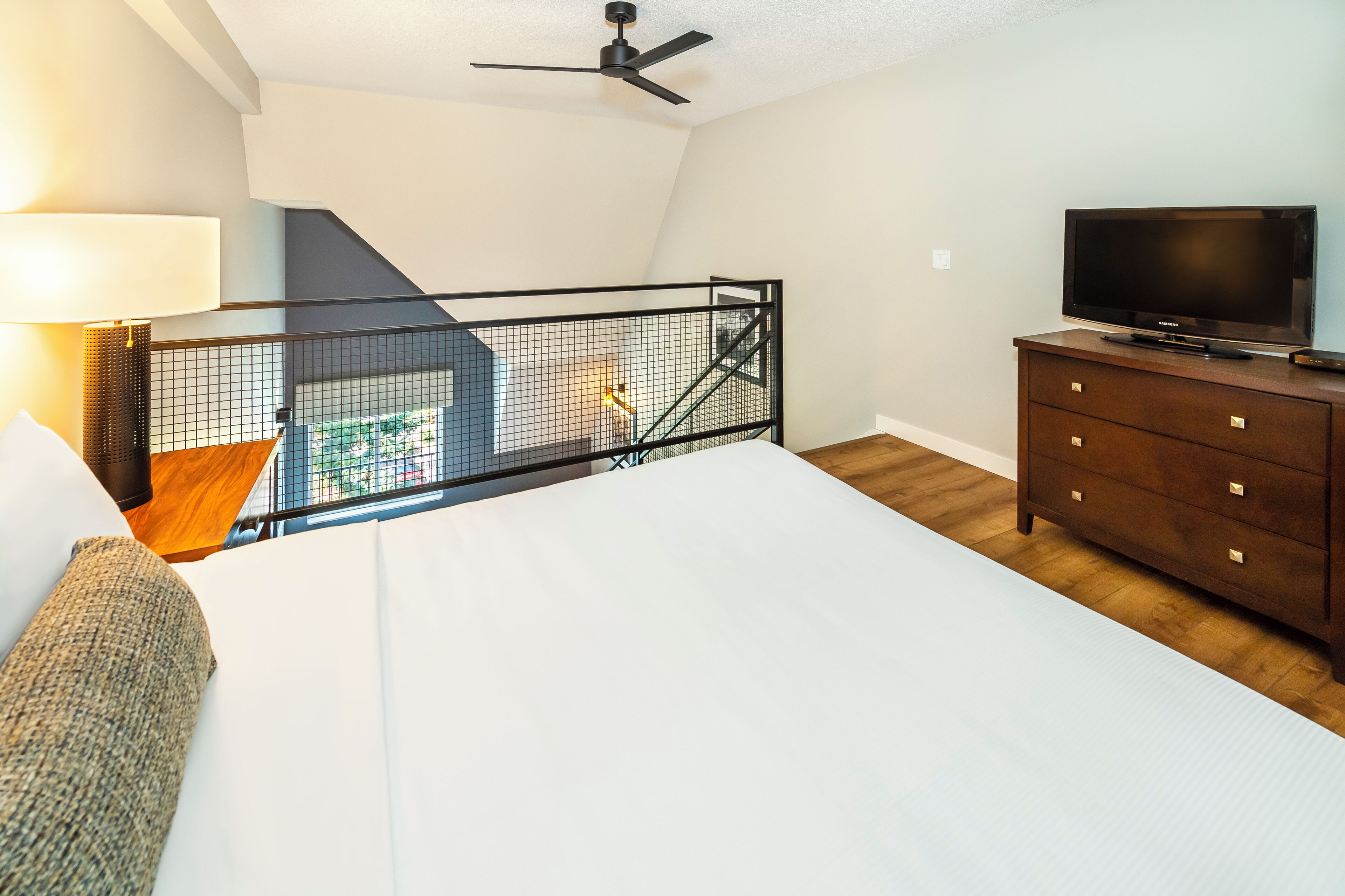 A bedroom with a bed, a TV on a dresser, a ceiling fan, and a view of a balcony with a railing.