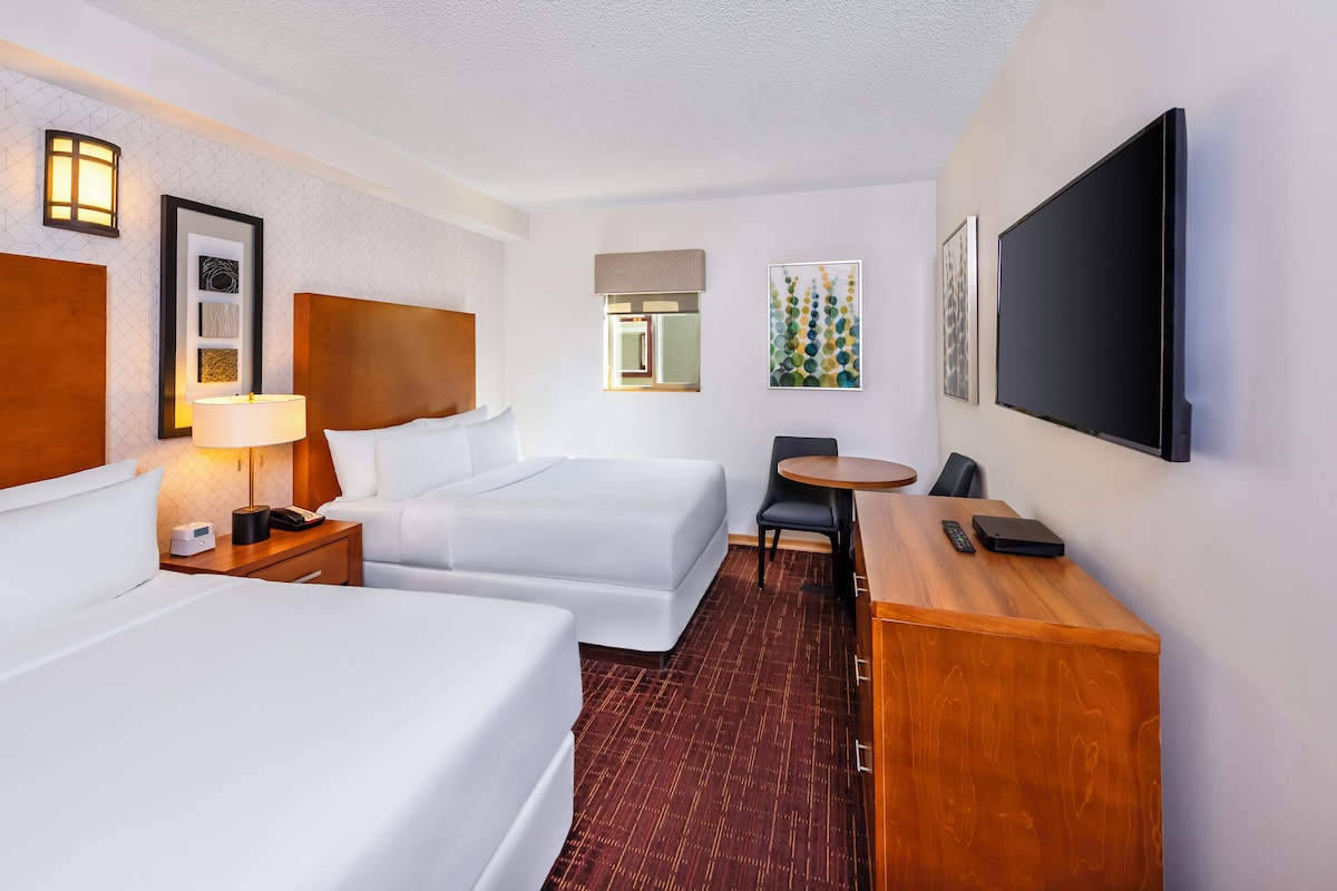A hotel room with two beds, a desk, a chair, a TV, and a lamp.