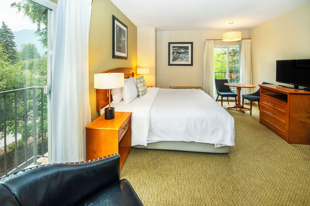 A hotel room with a large bed, a desk with a television, a chair, and a balcony with a view of trees and mountains.