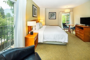 A hotel room with a large bed, a desk with a television, a chair, and a balcony with a view of trees and mountains.