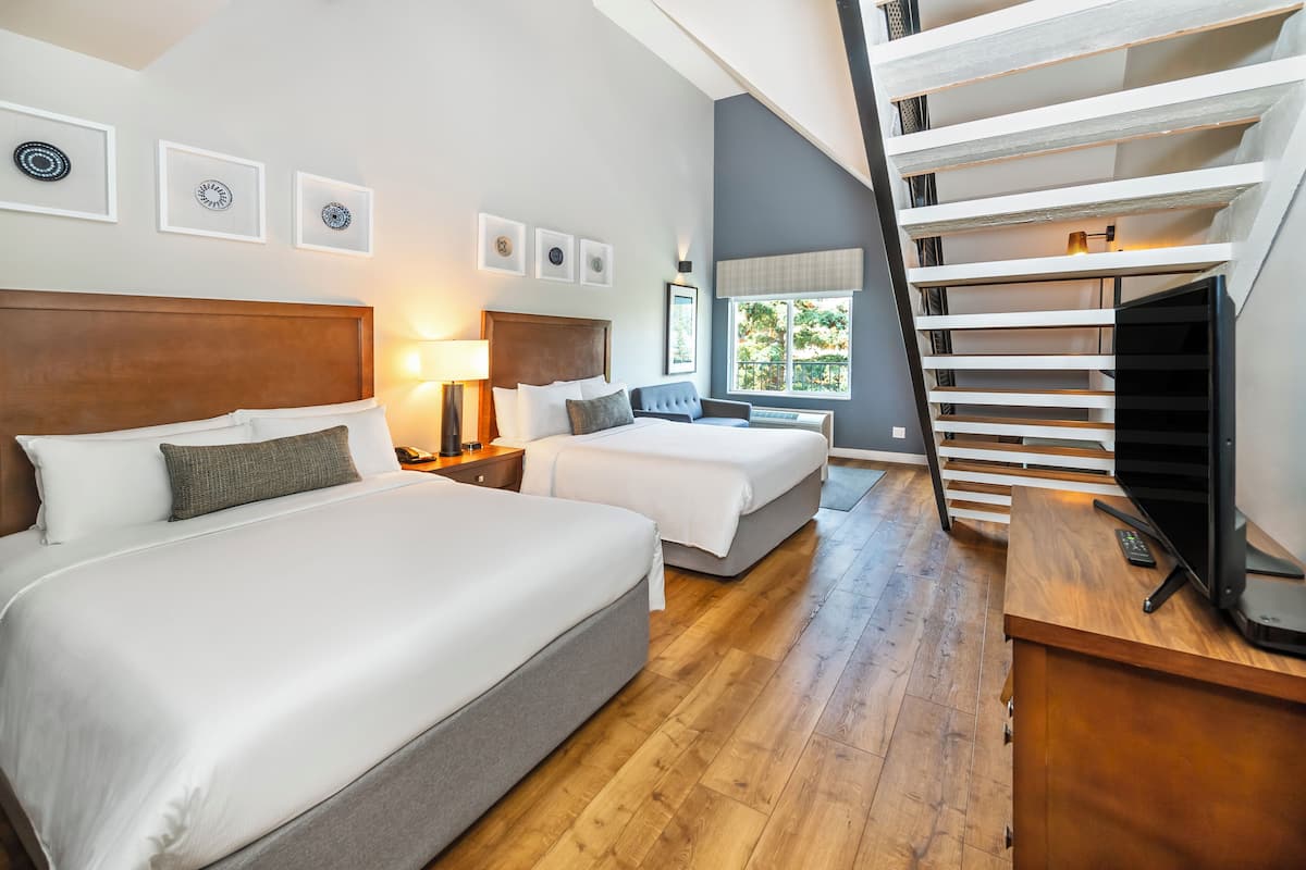 A hotel room with two beds, a wooden headboard, a nightstand with a lamp, a TV, and a staircase with a railing.