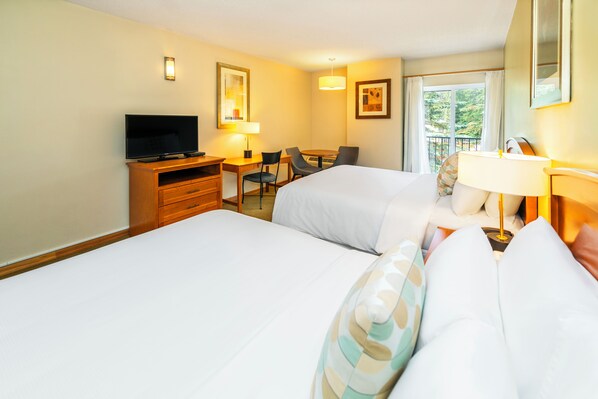 A hotel room with two beds, a desk with a TV, a chair, and a window with a view of greenery.