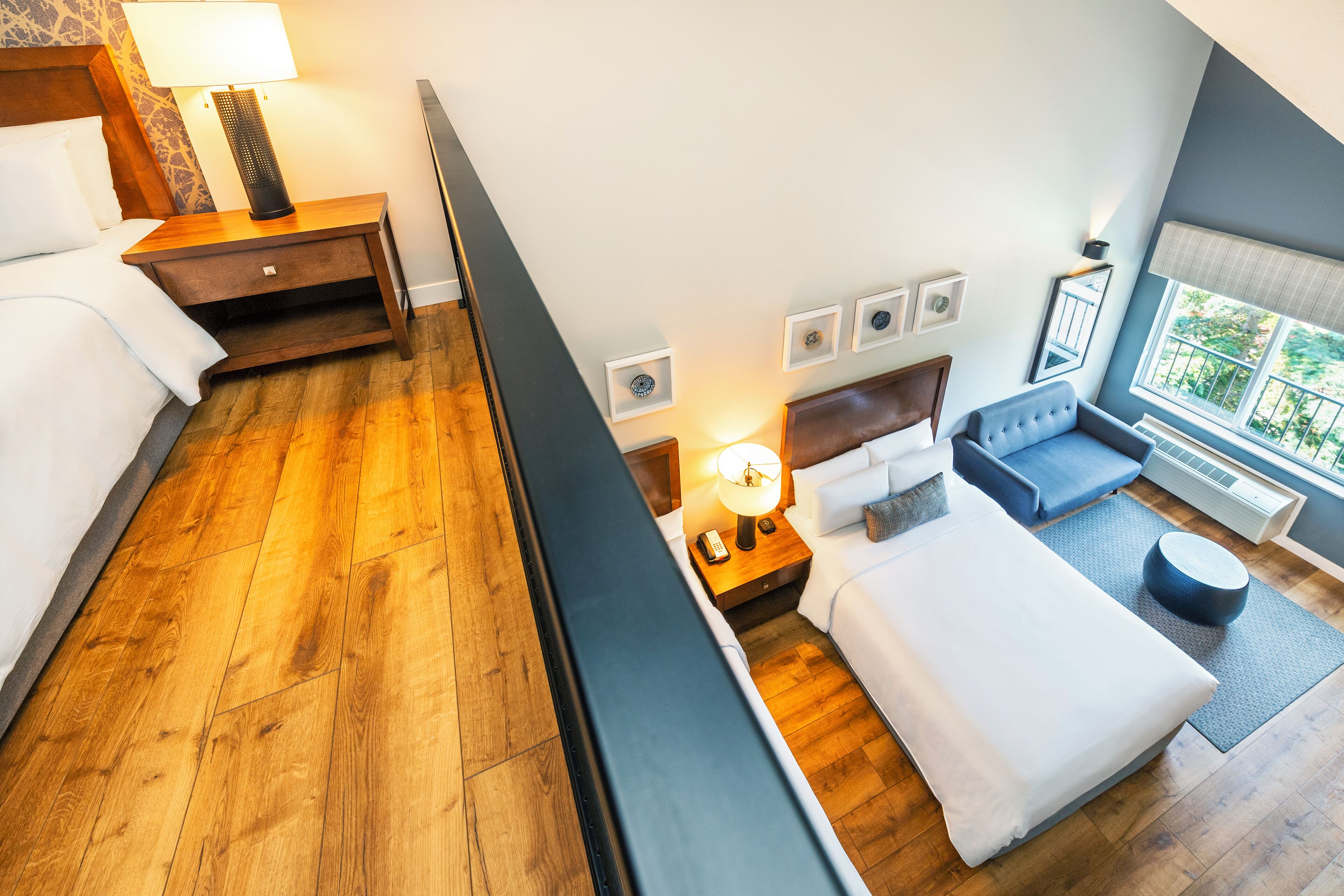 A hotel room with a wooden floor, a bed, a sofa, a small table with a lamp, and a window with a view of greenery.