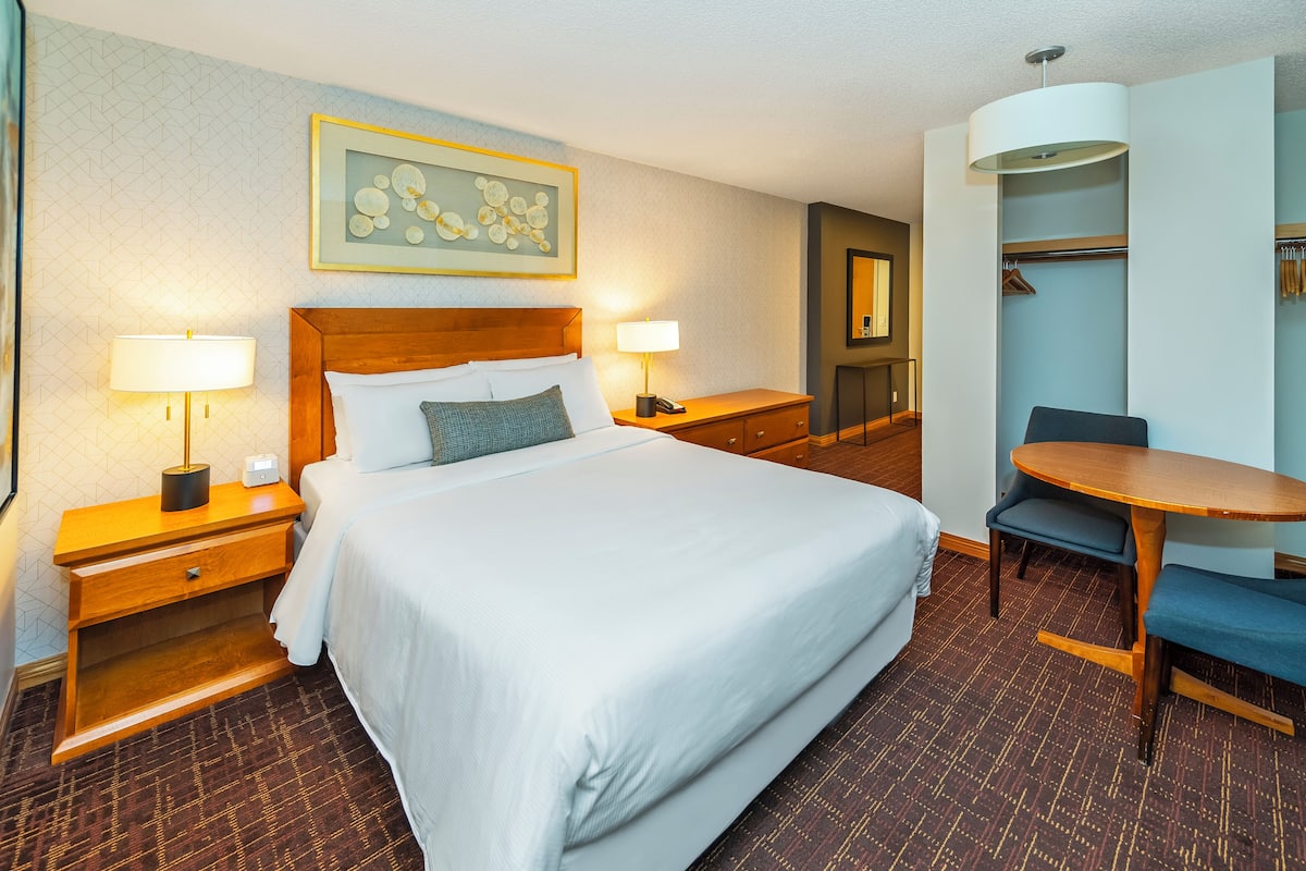 A hotel room with a large bed, bedside tables with lamps, a desk with a chair, a wardrobe, and a mirror.