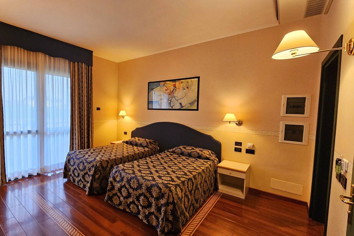 A hotel room with two beds, a large window with curtains, a painting on the wall, and a nightstand with a lamp.