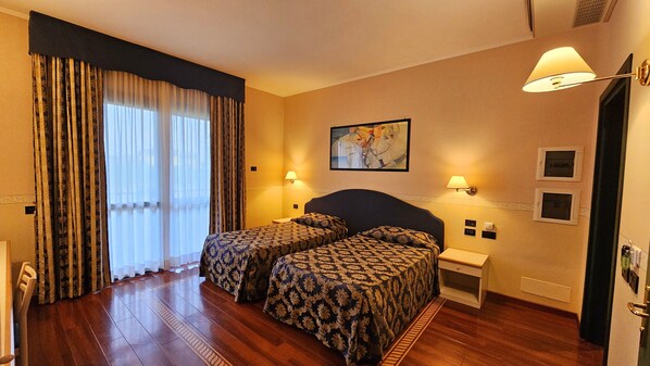 A hotel room with two beds, a large window with curtains, a painting on the wall, and a nightstand with a lamp.
