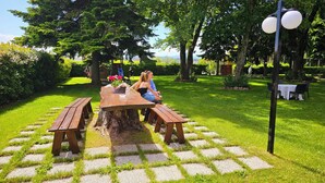A picnic table set on a stone pathway surrounded by lush greenery, with a playground in the background. - Ai Tigli (Langhirano)