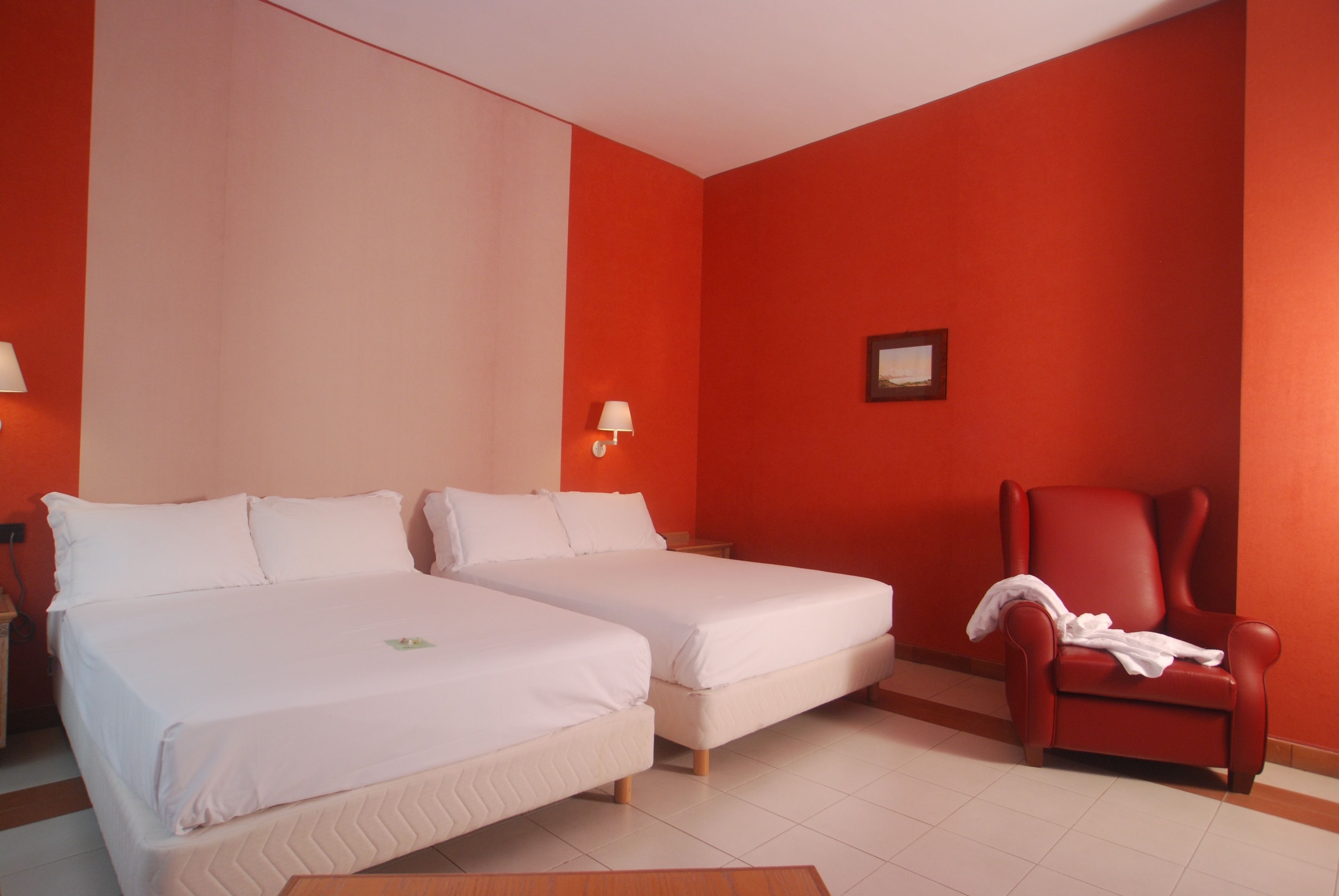 A hotel room with two beds, a red armchair, and a wall-mounted lamp.