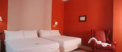 A hotel room with two beds, a red armchair, and a wall-mounted lamp.