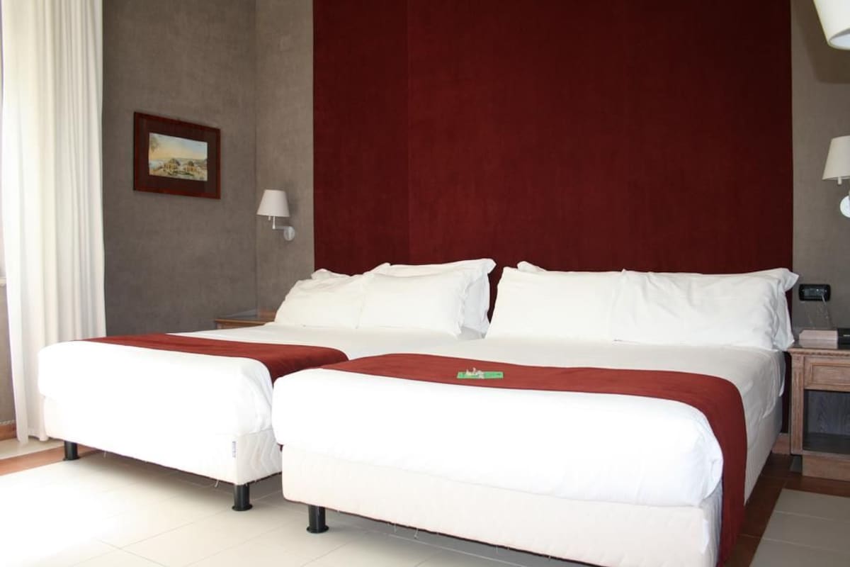 Una cama doble con ropa de cama blanca y un toque rojo, una mesita de noche con una lámpara y un cuadro en la pared.