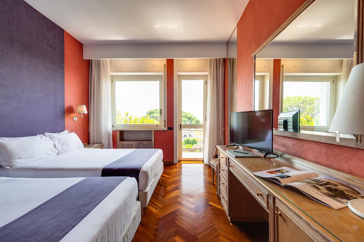 A hotel room with two beds, a TV, a desk with a lamp, and a large window with a view of greenery.