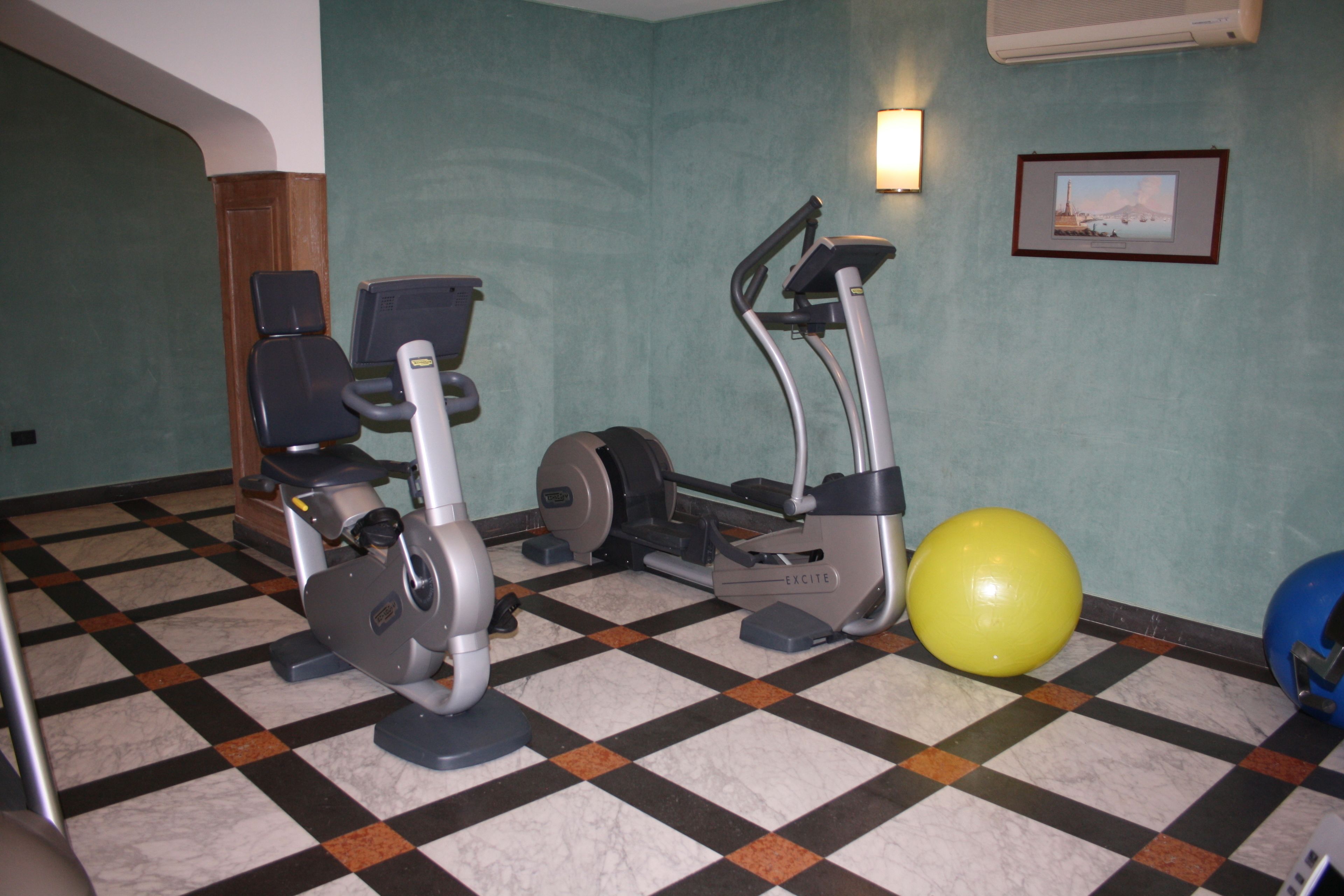 Un gimnasio con bicicletas estáticas y una pelota de ejercicios amarilla, suelo de cuadros y un cuadro en la pared.