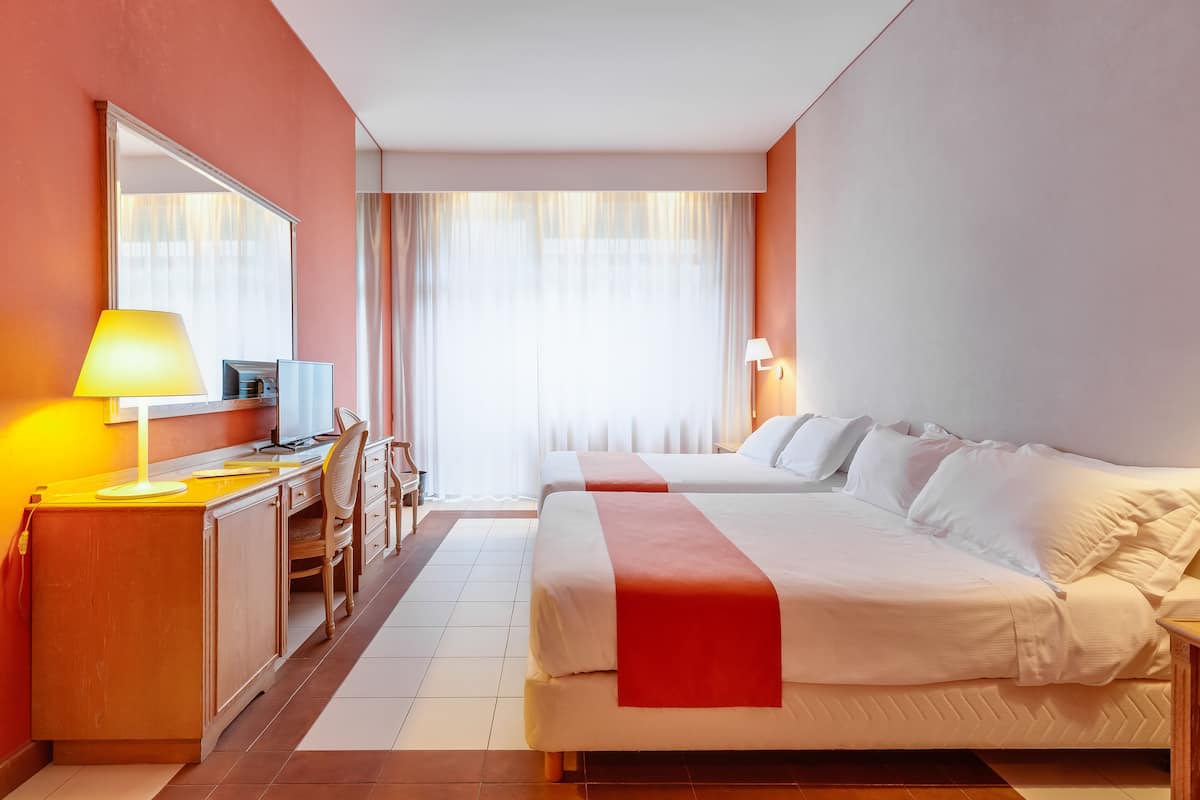 A hotel room with two beds, a desk with a lamp, a chair, and a large window with curtains.