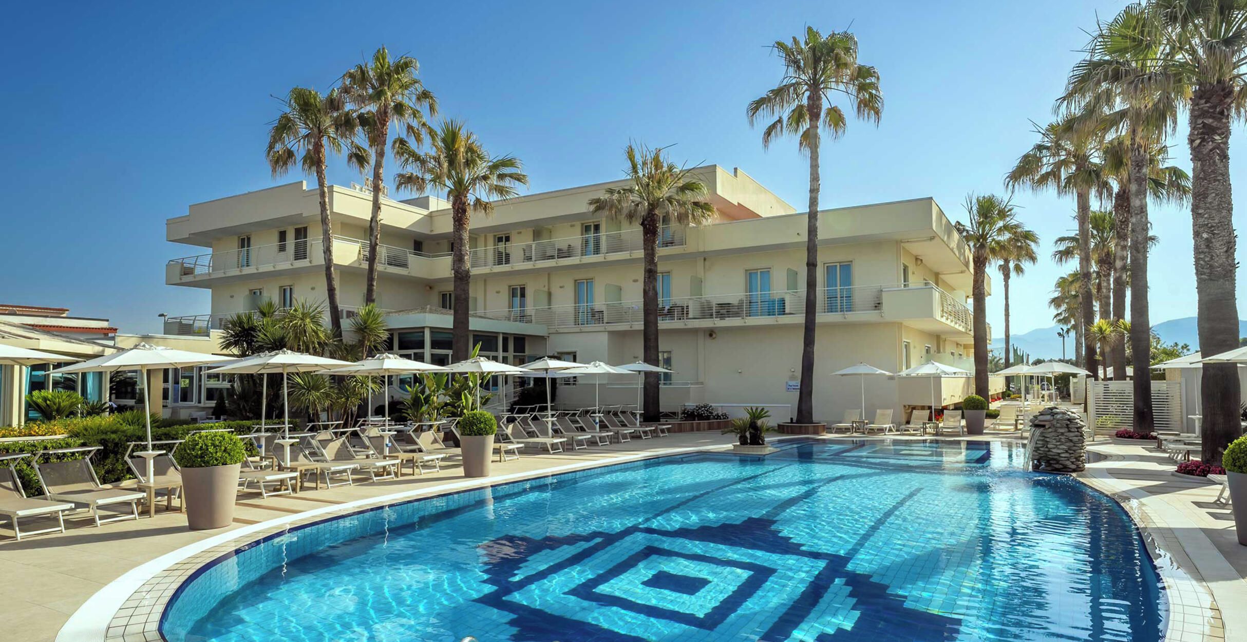 A swimming pool with lounge chairs and umbrellas, surrounded by palm trees and a multi-story building.