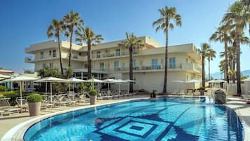 A swimming pool with lounge chairs and umbrellas, surrounded by palm trees and a multi-story building.