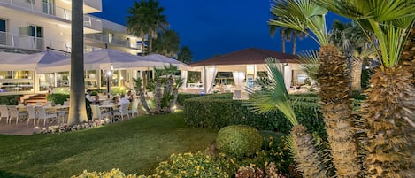 An outdoor dining area with tables and chairs, surrounded by palm trees and well-maintained hedges.