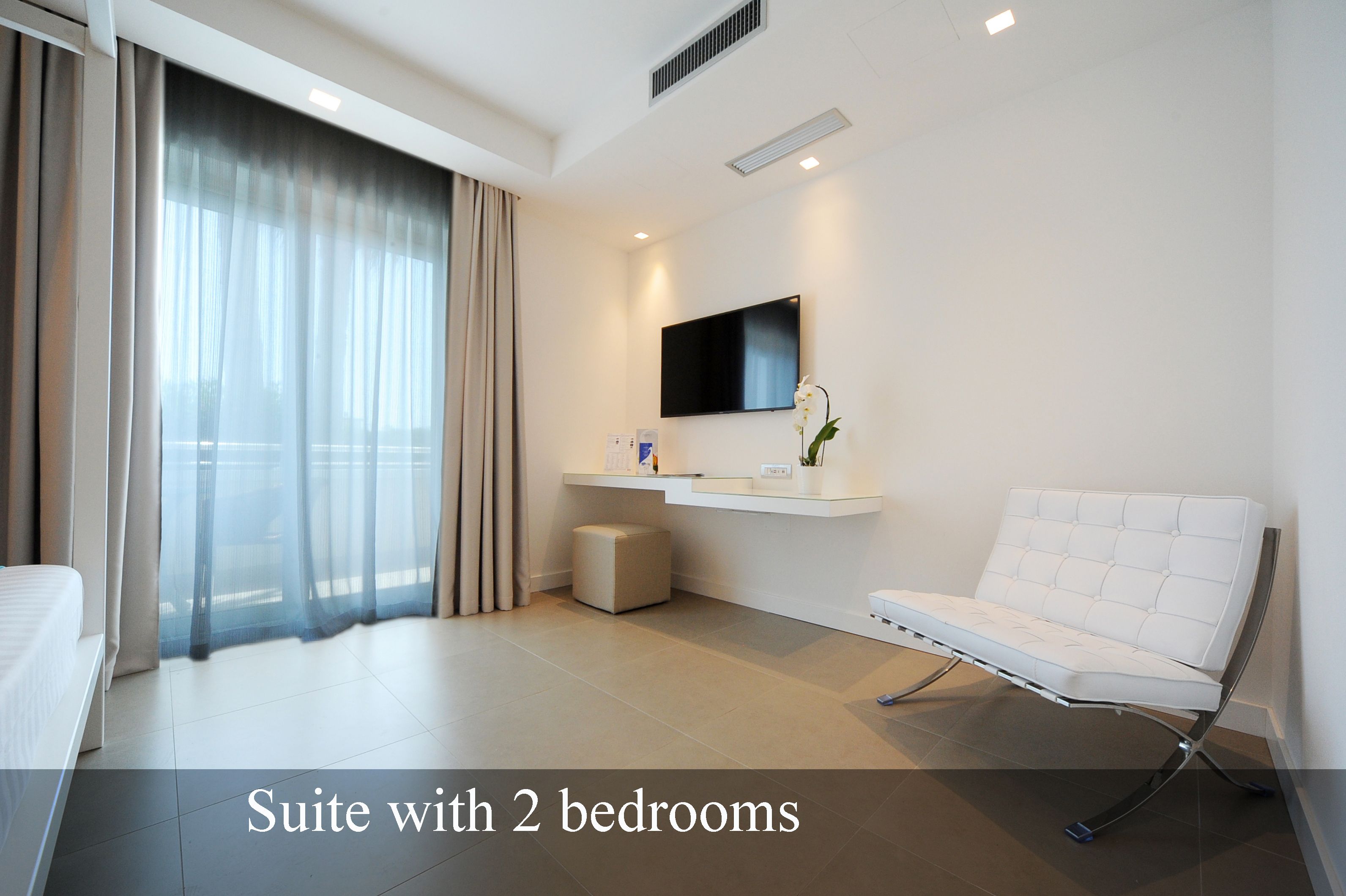 Suite d'hotel moderna amb dues habitacions, televisió de pantalla plana, sofà blanc i una finestra gran amb cortines transparents.