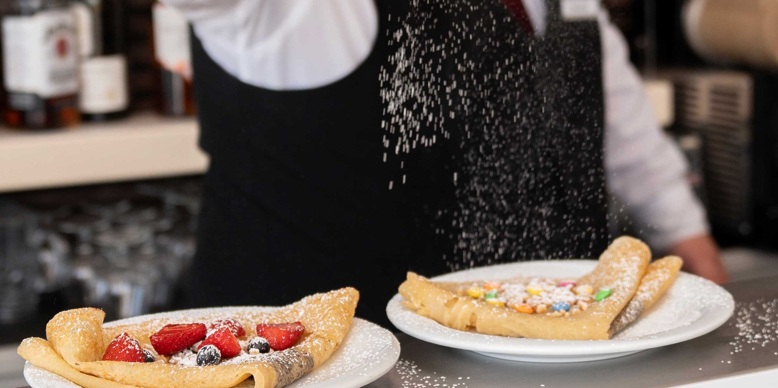Une personne saupoudre du sucre glace sur une assiette de dessert contenant une crêpe et des fraises.
