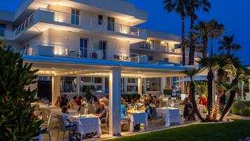 Un hotel de varios pisos con zonas de comedor al aire libre, palmeras y un porche cubierto.