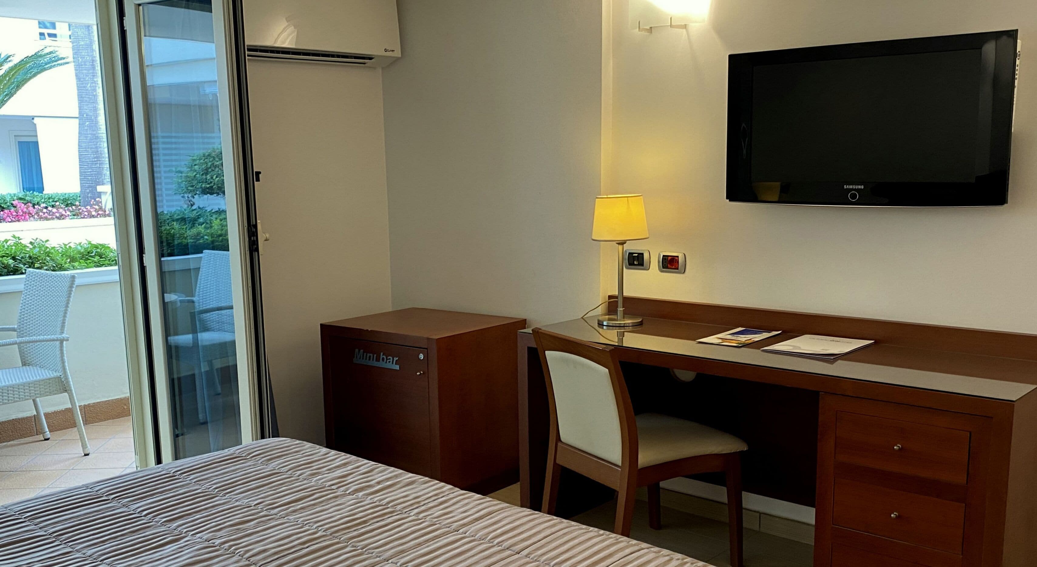 A hotel room with a bed, a desk, a chair, a minibar, a TV, and a window with a view of greenery.