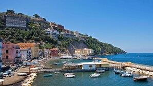 A coastal town with buildings on a hillside, a pier with boats, and a clear blue sea. - Hotel Olimpico (Pontecagnano Faiano)