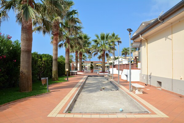 A courtyard with a croquet field, palm trees, and a building with a balcony. - Hotel Olimpico (Pontecagnano Faiano)