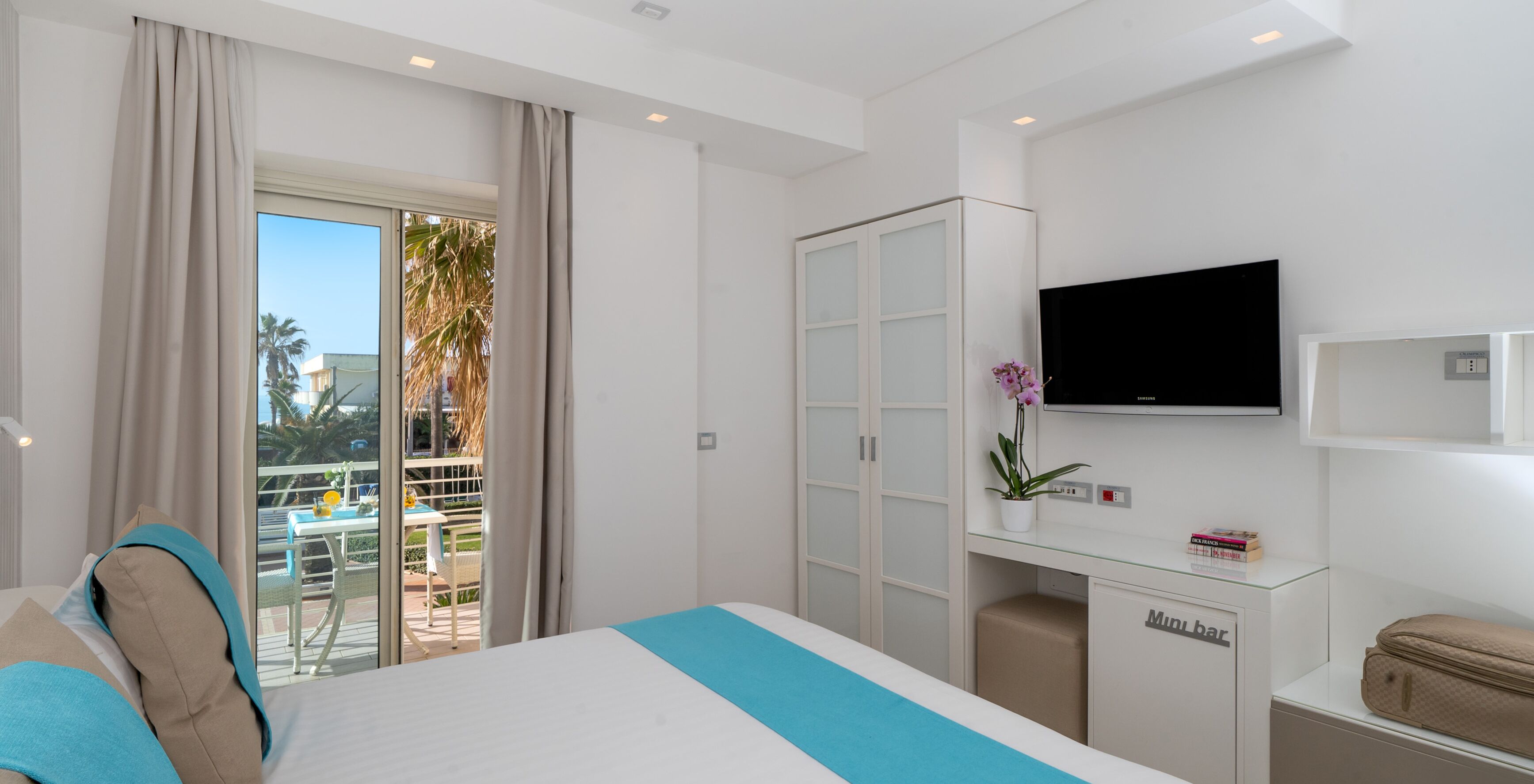 A hotel room with a bed, a minibar, a television, and a balcony with a view of palm trees.