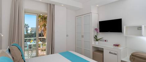 A hotel room with a bed, a minibar, a television, and a balcony with a view of palm trees.