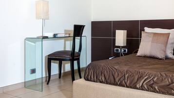 Habitación de hotel moderna con escritorio de vidrio, sillón de cuero, cama marrón con cubrecama texturizado y cabecera de madera oscura.