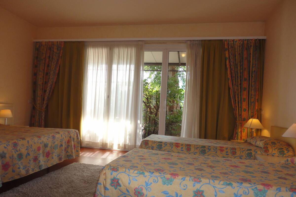 Ein Hotelzimmer mit zwei Betten, einem großen Fenster inklusive Vorhängen und Blick auf Grün.