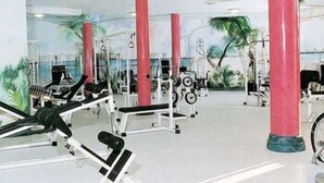 Et fitnesscenter med forskellig træningsudstyr, herunder vægtmaskiner og bænke, samt store vinduer med udsigt til palmer.