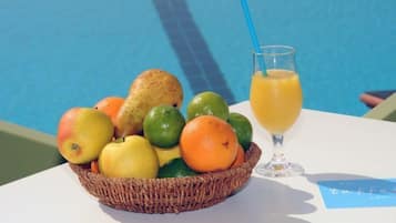 Een fruitmand met appels, sinaasappels en limoenen naast een glas jus d'orange bij een zwembad.